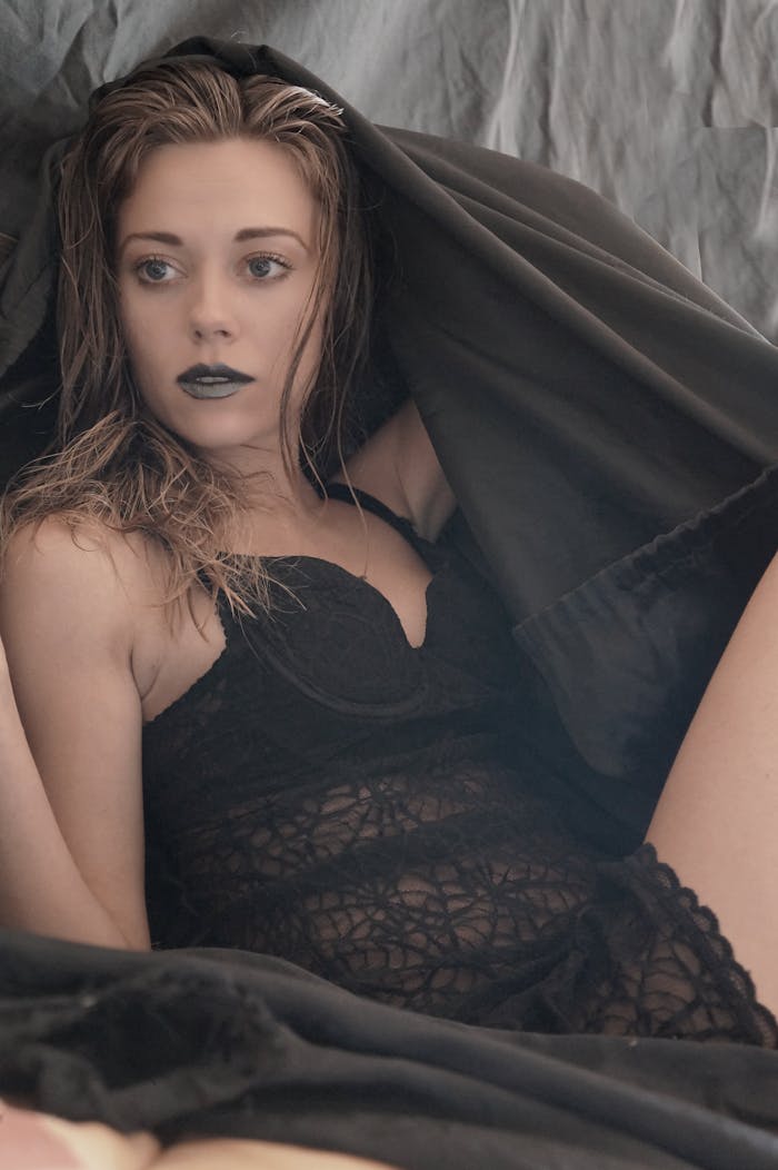 A model poses elegantly in black lace lingerie under a draped fabric, exuding a moody and artistic vibe.