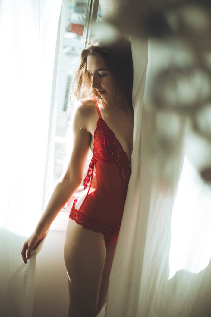 Artistic portrait of a woman in red lingerie standing by a bright window.
