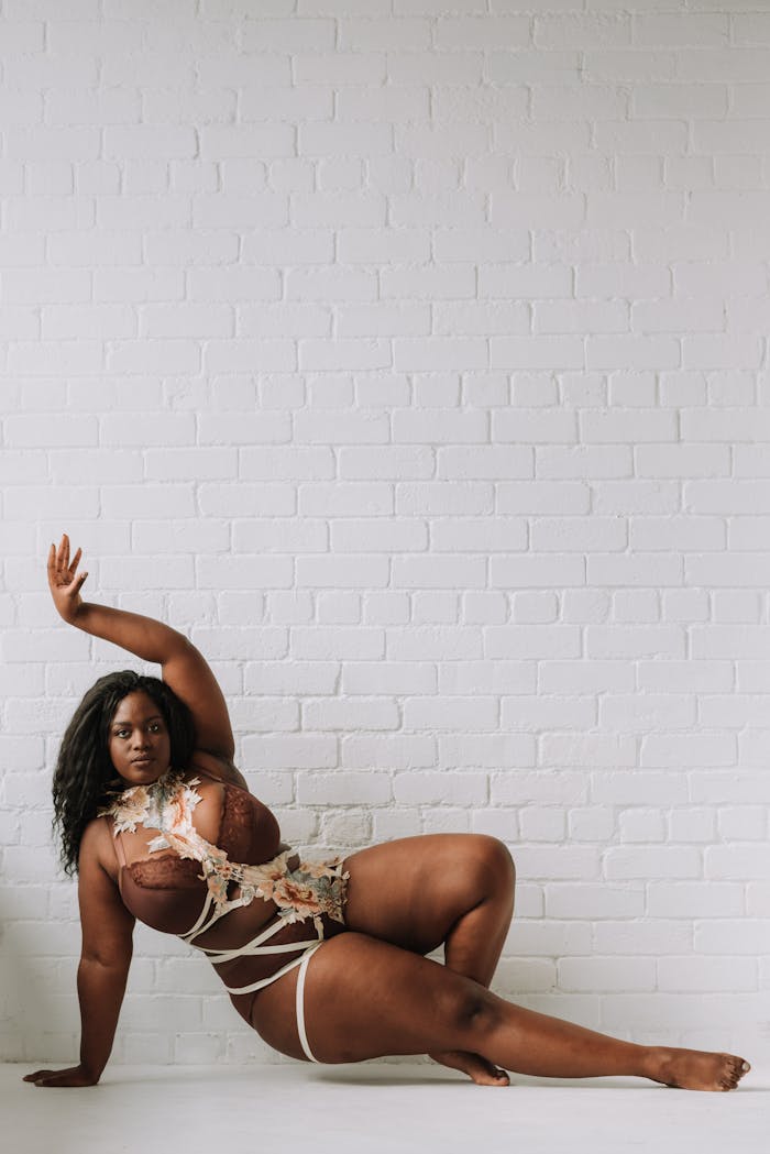 Confident African American woman in creative lingerie pose against a white brick wall, celebrating body positivity.